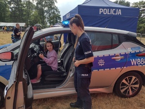 Zdjęcie kolorowe. W radiowozie siedzi na miejscu kierowcy dziewczynka. Obok radiowozu przy otwartych drzwiach stoi policjantka