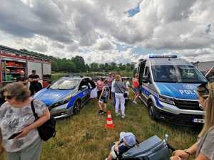 Zdjęcie kolorowe. Na terenie zielonym stoją dwa zaparkowane policyjne radiowozy oraz wóz strażacki. Wokół stoją osoby czekające na wejście do środka pojazdów