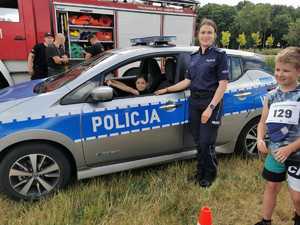 Zdjęcie kolorowe. Policjantka stoi na tle radiowozu w środku na miejscu kierowcy siedzi dziewczynka