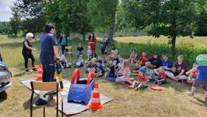 Zdjęcie kolorowe. Grupa dzieci siedzi na trawie. Przed nimi stoi policjantka, która trzyma piankowy sygnalizator świetlny, za nią stoi zaparkowany radiowóz