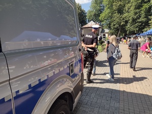 Zdjęcie kolorowe. Z lewej strony część boczna policyjnego busa. Za nim stoi umundurowany policjant, który rozmawia z mężczyzną