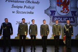 Zdjęcie kolorowe. Na scenie stoi czterech policjantów. Z lewej strony szeregu stoi komendant powiatowy z prawej komendant wojewódzki
