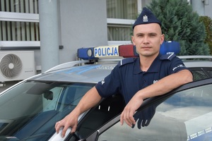 Zdjęcie kolorowe. Umundurowany policjant na tle radiowozu.