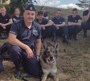 Zdjęcie. Na zdjęciu przewodnik psa służbowego wraz z psem Kalą. W tle siedzą uczniowie klas policyjnych w mundurach.