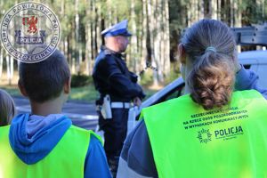 Zdjęcie kolorowe. Na pierwszym planie stoją odwrócone plecami dzieci w kamizelkach odblaskowych. W tle widać policjanta