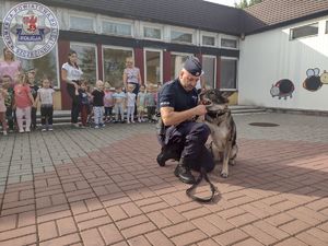 Zdjęcie kolorowe. Policjant kuca przy swoim psie. W tle widać dzieci