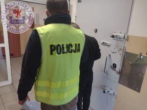 Zdjęcie kolorowe. Policjant ubrany w kamizelkę odblaskową odwrócony jest plecami, obok tyłem stoi mężczyzna ubrany na czarno