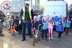 Zdjęcie kolorowe. Policjant stoi w towarzystwie dzieci. Jedno z nich trzyma koło na rączce tzw.rolkę służącą do pomiaru np. drogi hamowania