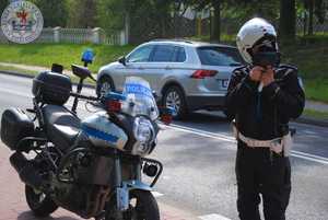 Zdjęcie kolorowe. Policjant mierzy prędkość, za nim stoi motocykl