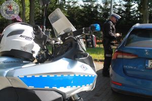 Zdjęcie kolorowe. Na pierwszym planie fragment policyjnego motocyklu w tle kontrolujący pojazd policjant