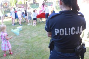 Zdjęcie kolorowe. Policjantka odwrócona plecami patrzy w kierunku bawiących się dzieci