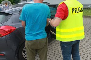 Zdjęcie kolorowe. Zatrzymanie. Mężczyzna w niebieskiej koszulce stoi obok policjanta w kamizelce odblaskowej z napisem policja