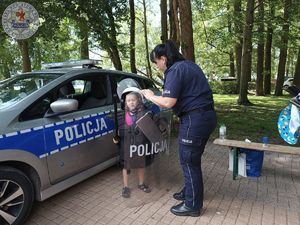 Zdjęcie kolorowe. Policjantka pomaga przytrzymać tarczę dziecku ubranemu w strój podwyższonego ryzyka