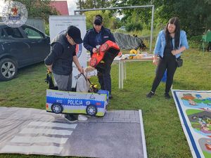 Zdjęcie kolorowe. Policjantka uczy dzieci przechodzenia przez przejście dla pieszych z wykorzystaniem piankowych samochodzików na szelkach i maty wyglądającej jak droga