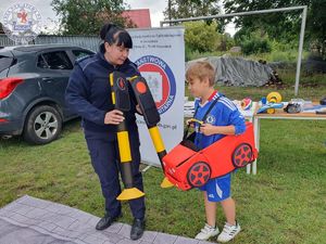 Zdjęcie kolorowe. Policjantka uczy dzieci przechodzenia przez przejście dla pieszych z wykorzystaniem piankowych samochodzików na szelkach i maty wyglądającej jak droga