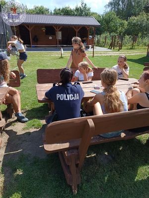 Zdjęcie kolorowe. Policjantka siedzi na zewnątrz przy stole z dziećmi. W tle budynki gospodarcze.