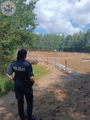 Zdjęcie kolorowe. Policjantka odwrócona plecami obserwuje dzieci jeżdżące na koniach