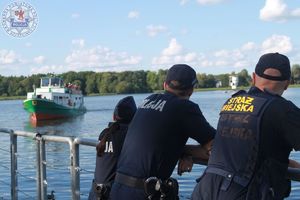 Zdjęcie kolorowe. Z lewej strony kadru widać tramwaj wodny na jeziorze. Z prawej oparty o balustradę pomostu strażnik miejski i para policjantów spoglądających na tramwaj