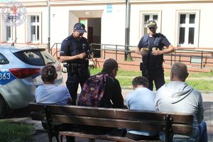 Zdjęcie kolorowe. Policjant ze strażnikiem miejskim legitymuje osoby siedzące na ławce