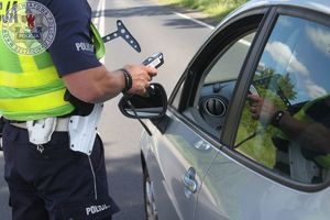 Zdjęcie kolorowe. Policjant stoi przy samochodzie od strony kierowcy. W dłoni trzyma urządzenie do badania stanu trzeźwości