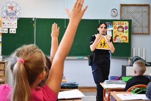 Zdjęcie kolorowe. Na pierwszym planie dziewczynka z podniesioną ręką w tle policjantka, która trzyma znak drogowy&quot;uwaga dzieci&quot;