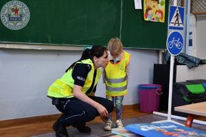 Zdjęcie kolorowe. Policjantka w kamizelce odblaskowej kuca przy dziewczynce również w kamizelce odblaskowej