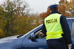 Zdjęcie kolorowe. Policjant bada kierującego, który siedzi za kierownicą granatowego samochodu