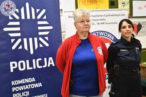 Zdjęcie kolorowe. Policjantka i promotorka zdrowia pozują do zdjęcie na tle logotypów