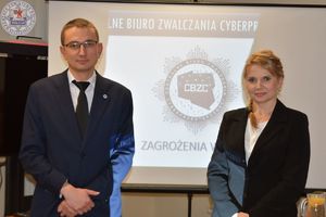 Zdjęcie kolorowe. Kobieta i mężczyzna stoją na tle białego ekranu na którym wyświetlone jest logo Centralnego biura Zwalczania Cyberprzestępczości