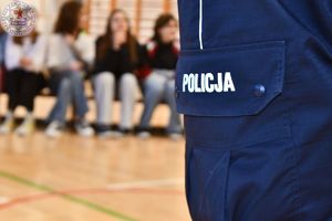 Zdjęcie kolorowe. Zbliżenie napisu policja na nogawce policyjnego munduru, w tle dzieci siedzące na ławeczce