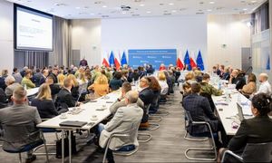 Zdjęcie kolorowe. Przy długich stołach siedzą ludzie. W tle widać flagi Polski i UE