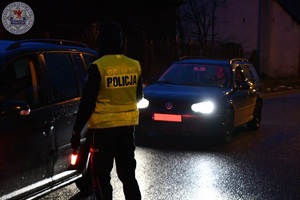 Zdjęcie kolorowe. Zdjęcie zrobione w nocy. Policjant kontroluje kierującego na zawartość alkoholu w wydychanym powietrzu