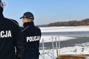 Zdjęcie kolorowe. Policjanci kontrolują linię brzegową przy zamarzniętym jeziorze