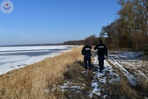 Zdjęcie kolorowe. Policjanci kontrolują linię brzegową przy zamarzniętym jeziorze