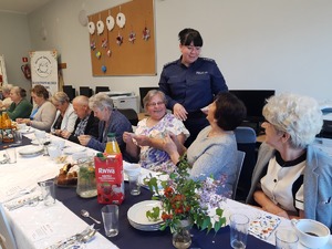 Zdjęcie kolorowe. Sala wypełniona osobami zasiadającymi przy zastawionych stołach. Przed nimi stoi policjantka