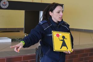 Zdjęcie kolorowe. Policjantka trzyma znak drogowy. Na żółtym tle dziewczynka trzyma czerwonnego cukierka na patyku