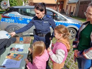 Zdjęcie kolorowe. Policjantka obok pojazdu służbowego w obecności dzieci