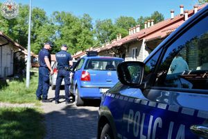 Zdjęcie kolorowe. Policjanci kontrolują pojazd koloru niebieskiego. Widoczny fragment policyjnego radiowozu