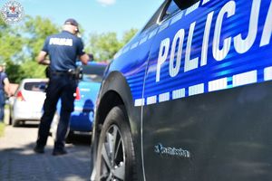 Zdjęcie kolorowe. Policjanci kontrolują pojazd koloru niebieskiego. Widoczny fragment policyjnego radiowozu