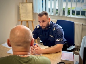 Zdjęcie kolorowe. Łysy mężczyzna siedzi tyłem, przed nim przy biurku policjant