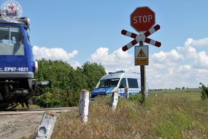 Zdjęcie kolorowe. Przejazd kolejowy z uchwyconym fragmentem pociągu. Za przejazdem policyjny radiowóz