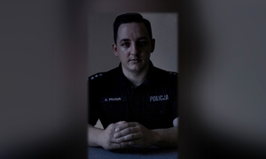 Zdjęcie kolorowe. Zdjęcie policjanta siedzącego przy biurku. Zdjęcie w ciemnej i mrocznej kolorystyce.