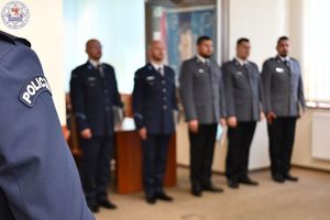 Zdjęcie kolorowe. Naszywka z napisem policja na ramieniu policyjnego munduru, w tle policjanci