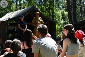 Zdjęcie. Harcerze siedzą na polanie. Przed nimi strażak i policjantka omawiają kwestie bezpieczeństwa