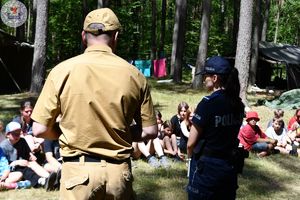 Zdjęcie. Harcerze siedzą na polanie. Przed nimi strażak i policjantka omawiają kwestie bezpieczeństwa