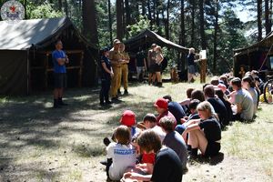 Zdjęcie. Harcerze siedzą na polanie. Przed nimi strażak i policjantka omawiają kwestie bezpieczeństwa