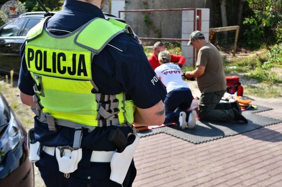 Zdjęcie kolorowe. Policjant ruchu drogowego, w tle stanowisko do ćwiczeń z pierwszej pomocy