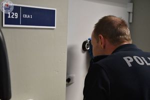 Zdjęcie kolorowe. Policjant przez wizjer obserwuje co się dzieje w celi