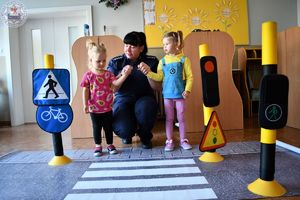 Zdjęcie kolorowe. Dzieci w sali ćwiczą przechodzenie przez przejście dla pieszych. Policjantka kuca i trzyma dwie dziewczynki. Przed nimi mata z przejściem dla pieszych