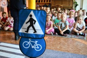 Zdjęcie kolorowe. Zbliżenie znaku drogowego przejście dla pieszych i droga dla rowerów. W tle siedzące na dywanie dzieci